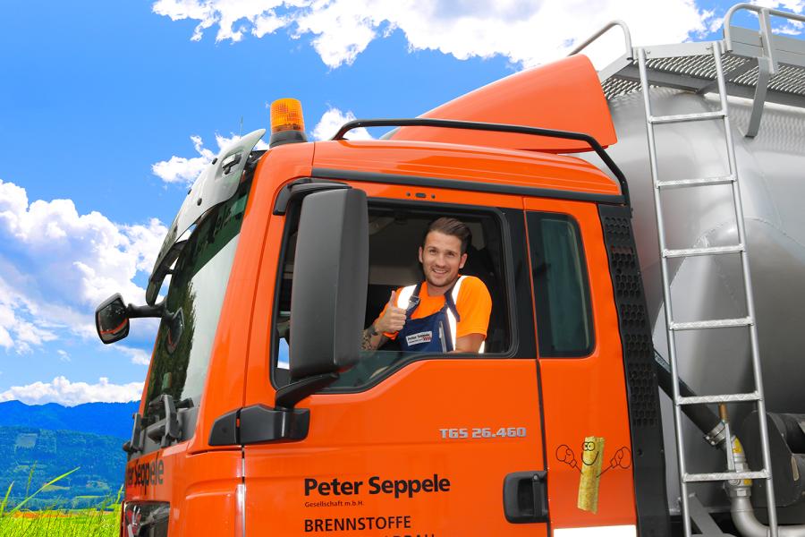 LKW Fahrer der Firma Seppele transportiert Heizinos Pellets | © Peter Seppele GmbH Ein freundlicher Lkw-Fahrer zeigt einen Daumen hoch aus seinem Lastwagenkabinenfenster. Der orangefarbene Lkw ist auf einer malerischen Strecke mit strahlend blauem Himmel unterwegs. Im Hintergrund sind grüne Wiesen und Berge zu sehen, was eine einladende Atmosphäre schafft.